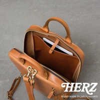 一個棕色皮革後背包，頂部提把和可拆卸肩帶，背包打開展示內部，一個平板電腦放入內袋，下方有 HERZ LOGO 和 HAND MADE IN JAPAN Since1973 字樣