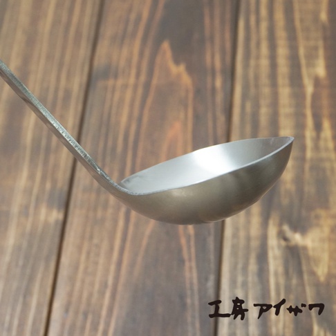 不鏽鋼萬用湯勺，勺柄和勺身均為銀色不鏽鋼材質，勺身呈弧形，背景為木紋桌面。