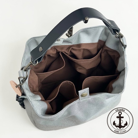 這是一款由神戶 clueto 品牌設計的 BALON mini 防潑水手提包，採俯視角度拍攝。包身外部為灰色防潑水材質，搭配黑色皮革提把與金屬扣環。包包內部設有多個深咖啡色內袋，具備良好的收納功能，右下方印有 clueto 的船錨品牌標誌。