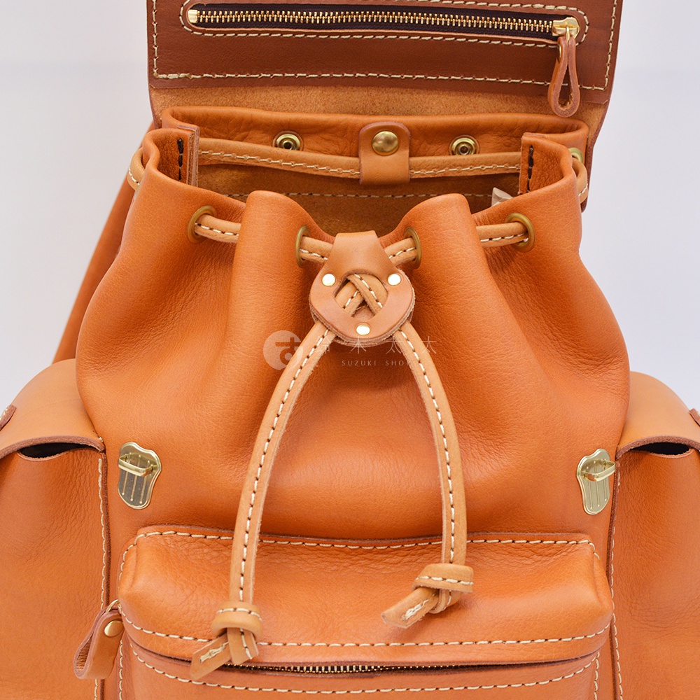 職人手作皮革復古風旅行後背包 R-65，展示部分為溫暖的棕褐色皮革，帶有抽繩開口設計，並有精緻的縫線細節與金屬扣件。