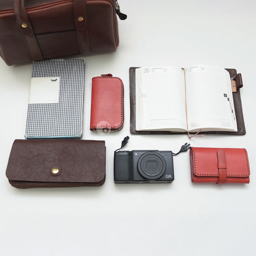 各種皮革配件，包含一個棕色波士頓包、一個格子筆記本、一個紅色零錢包、一本打開的日誌、一個皮夾、一台數位相機，以及一個紅色皮製卡夾。