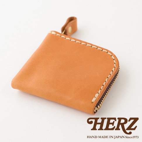 這是一款由日本品牌HERZ職人手工製作的駝色皮革零錢包，外觀採用極簡方形設計並搭配拉鍊封口，邊緣以粗縫線點綴，整體散發質感溫潤的皮革紋理，呈現細緻的手作工藝風格。