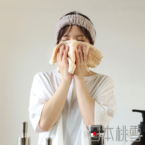 一位女子戴著毛絨髮帶，用一條淡黃色紋理的紗布洗臉巾擦拭臉部，背景是簡潔的白色牆壁，下方角落有寫著「日本桃雪」的文字。