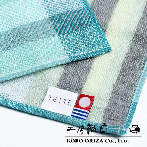 今治格紋信封式長方形手帕，採用棉質材質，交織著淺綠、灰、藍綠色調的格紋與條紋。手帕上有「TETE」品牌標籤，以及一個帶有紅、白、藍色條紋的紅色標誌。