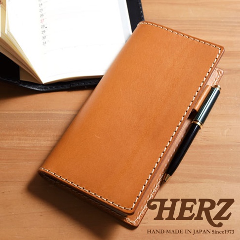 淺棕色皮革製成的職人手作輕便記事本書套，側面縫線清晰，右側夾有一支黑色金屬筆，書套左側露出部分打開的筆記本內頁，背景為木紋桌面。