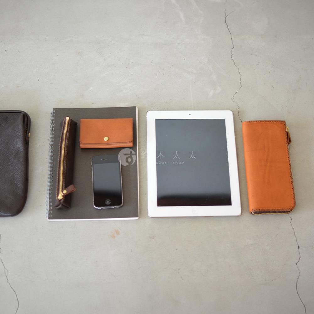 職人手作皮革Organ手拿包，展示一個深棕色皮革手拿包、一本筆記本、一個棕色皮革筆袋、一支手機、一台白色平板電腦和一個棕色皮革長夾，所有物品皆為皮革材質。