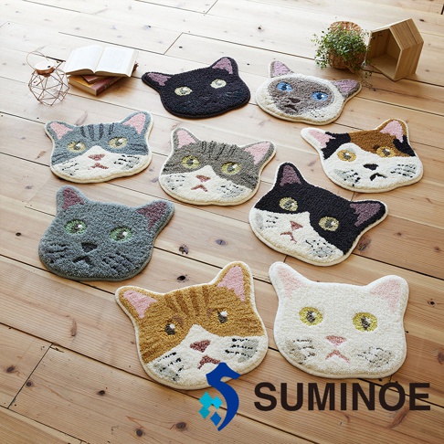 木質地板上陳列著八款SUMINOE品牌出品的NEKOKAO貓咪造型絨毛椅墊，這些椅墊呈現各式貓咪頭部特寫，包含黑貓、暹羅貓、三花貓、虎斑貓等多種花色，周邊裝飾著開書本、綠色盆栽與木質小屋模型。