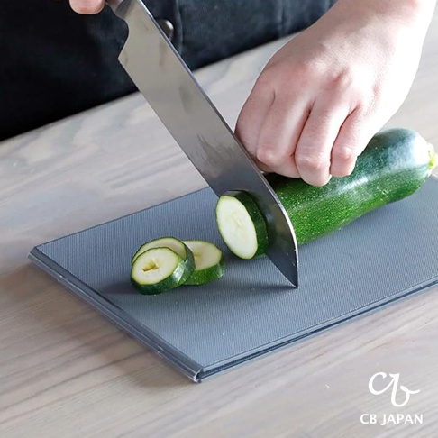CB JAPAN logo 標誌下的深灰色可折疊砧板上，一只手正用刀切一片櫛瓜。