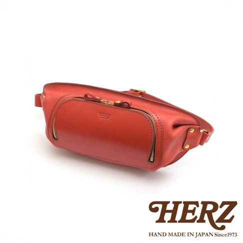 HERZ 職人手作紅色皮革拉鍊斜背包，正面有圓形皮標，金色拉鍊細節，產地為日本。