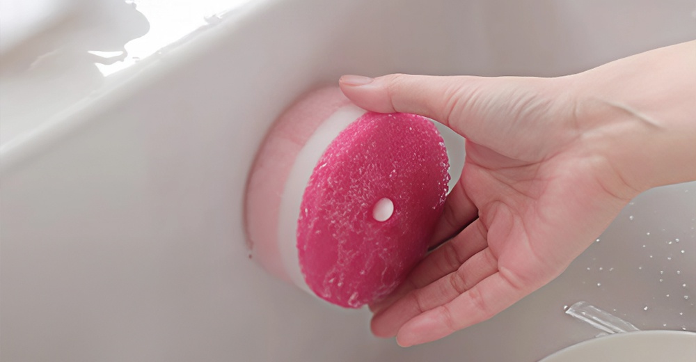 手持粉紅色圓形附吸盤洗碗海綿，海綿沾濕並有白色小水滴，海綿上有圓點。