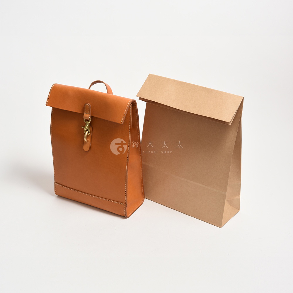 職人手作棕色皮革後背包，紙袋造型，配有金屬扣環和提把，旁為一個棕色紙袋。