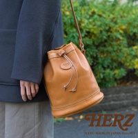 棕褐色皮革束口單肩包，包身圓潤，配有同色系皮革束繩及可調式細長肩帶，包身車縫白色明線，質感細膩。