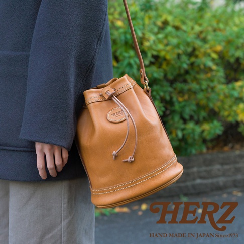 棕褐色皮革束口單肩包，包身圓潤，配有同色系皮革束繩及可調式細長肩帶，包身車縫白色明線，質感細膩。