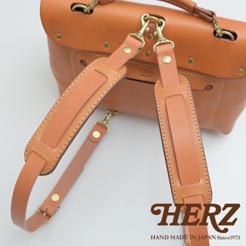 職人手作皮革肩墊，使用淺棕色皮革製作，附有黃銅色金屬扣環，可連接至包包。肩墊設計精緻，縫線清晰，展現職人細膩工藝。