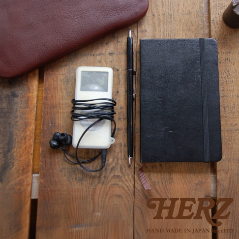 一疊皮革小物，包含一個酒紅色皮革封面、一個白色 iPod、一副黑色耳機、一支黑色鋼筆和一本黑色筆記本，擺放在木製桌面上。