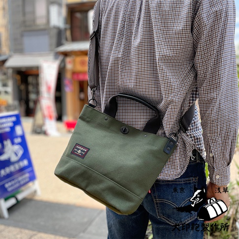 軍綠色帆布材質的兩用托特包，配有黑色提把和可拆卸肩帶，包蓋中央有黑色旋轉鎖扣，正面左側貼有方形品牌標籤，整體呈現簡約實用的風格。