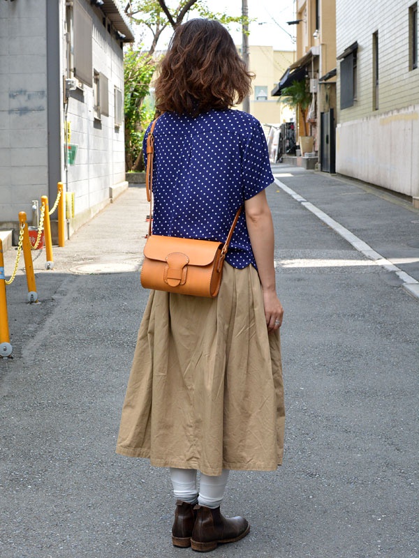一位女性身穿藍色圓點上衣與卡其色長裙，斜挎著一個 tan 色皮革馬鞍包，包上有寬版皮革扣帶。