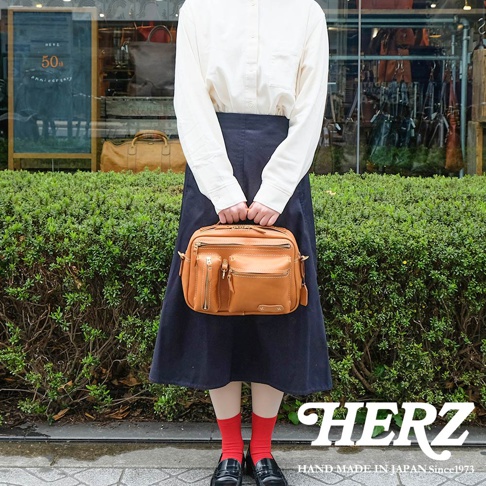 駝色職人手作皮革兩用多功能收納單肩包，正面有兩個拉鍊口袋和一個翻蓋口袋，搭配縫線細節，包身方正，適合日常攜帶。