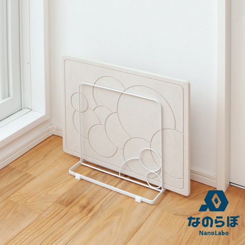 米白色珪藻土地墊，表面壓印有層疊圓形「泡泡」紋路，以白色金屬架支撐，置於木質地板上，背景為白色牆壁與窗戶。