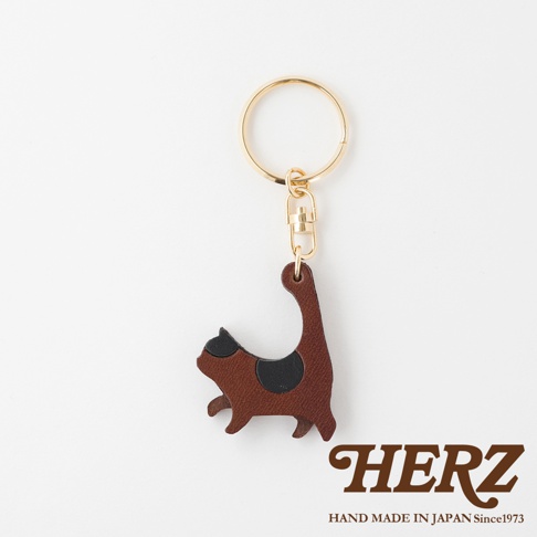咖啡色皮革製成的貓咪造型鑰匙圈，帶有黑色斑紋，搭配金色金屬掛環和連接扣，展現職人手作質感。