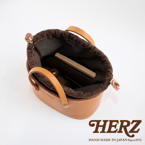 職人手作皮革束口手提包，淺棕色皮革，內襯為深棕色布料，束口處有繩結設計，提把為皮革材質，包內可見皮夾、手機及一本書。