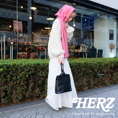 一位女性手持黑色皮革手提包，身穿白色針織衫、白色長裙與粉色頭巾，走在綠色灌木叢旁的街道上。