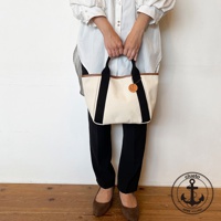 這款帆布手提托特包為米白色，配有黑色織帶和棕色皮革滾邊。包包上掛有一個圓形皮革吊牌，上面印有錨圖案。