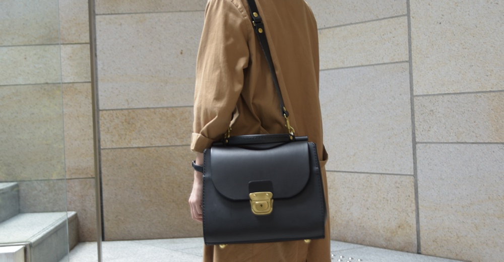 一名女子身著棕色外套，斜背著一個黑色皮革公事包，包包上有一個金色鎖扣。