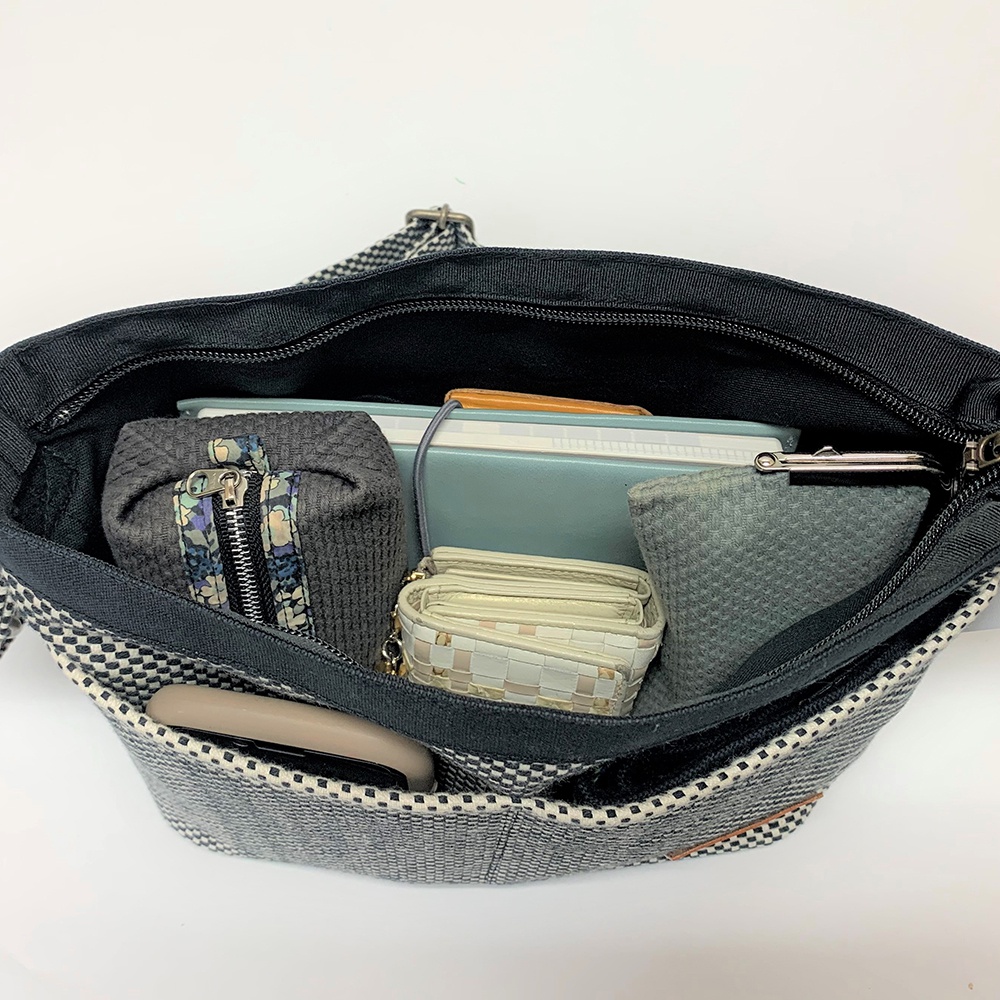 展示一個打開的三河木綿刺子織肩背包，內部裝有多樣物品，包括筆記本、手機、錢包、化妝包和零錢包，背包外部為黑白格紋織紋。