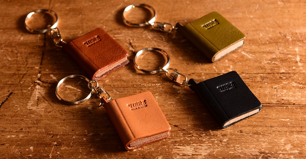 五款職人手作皮革書本造型鑰匙圈，包含棕色、橘色、橄欖綠及黑色，皆為皮革材質，附有金屬鑰匙圈。