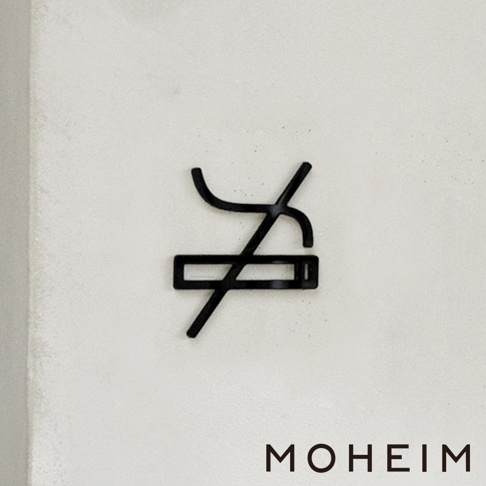 黑色金屬材質的「禁煙」標誌，由香菸圖案和斜線組成，標誌固定在白色紋理背景上。