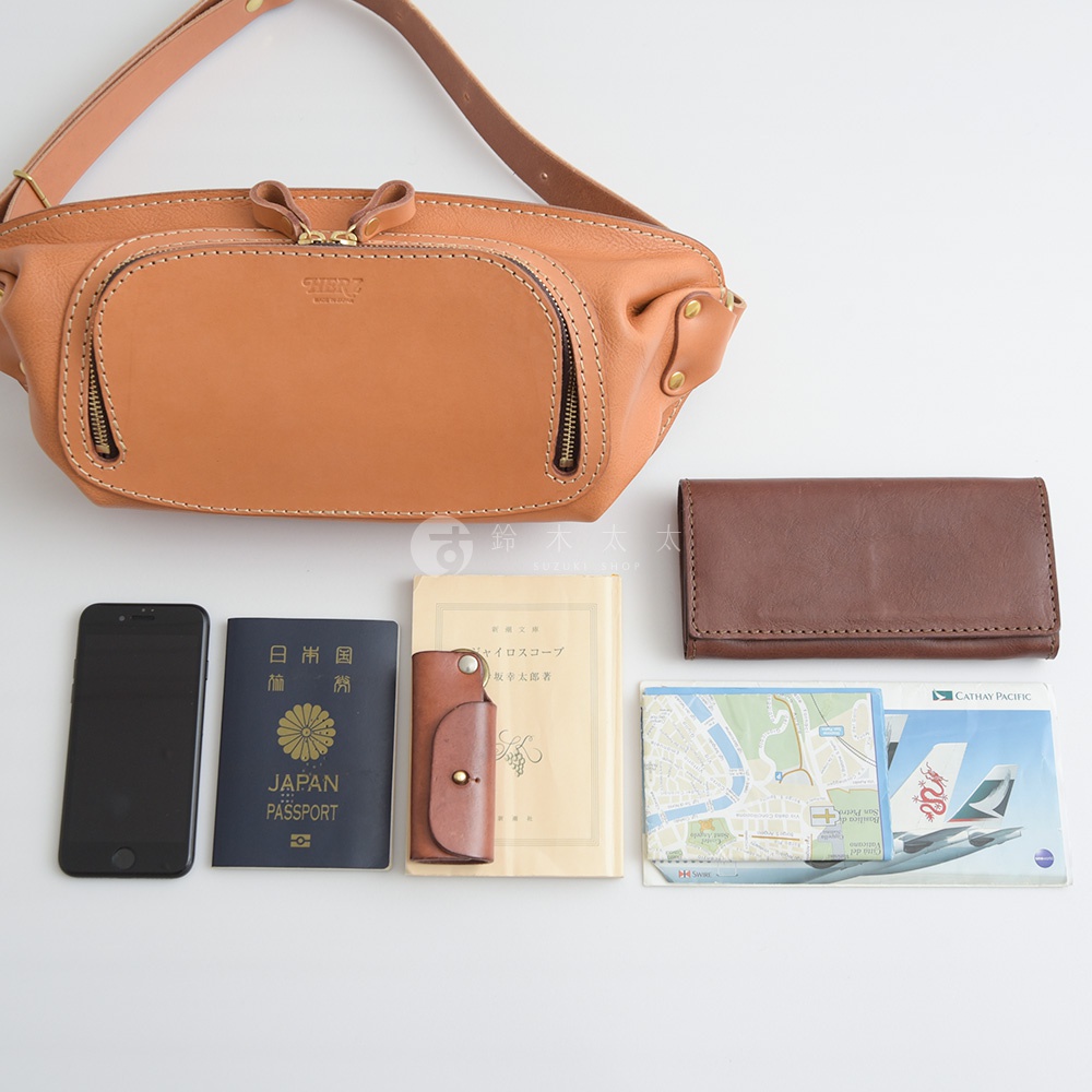 一個棕褐色的HERZ皮革斜背包，底部擺放著手機、日本護照、一個小皮夾、一本淺色書籍，以及一張地圖和一張國泰航空的機票。