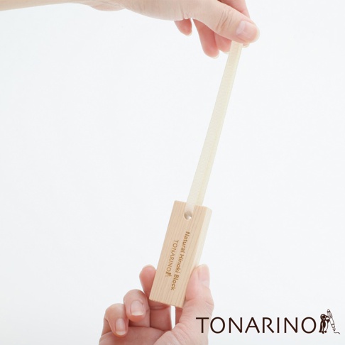 一隻手拿著一塊淺色天然檜木塊，檜木塊上印有「Natural Hinoki Block」及「TONARINO」字樣，一條米白色織帶穿過檜木塊上的孔洞，另一隻手握住織帶上方。