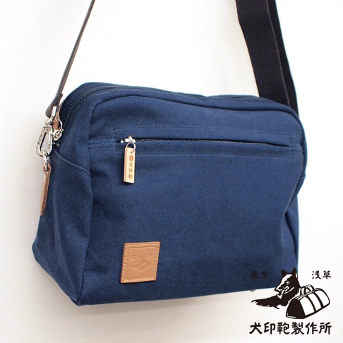 深藍色石洗帆布製成的旅行側背包，正面有一個拉鍊口袋，上面掛著一個木製吊飾，右下角有一個方形皮革標籤，上面印有圖案和文字。