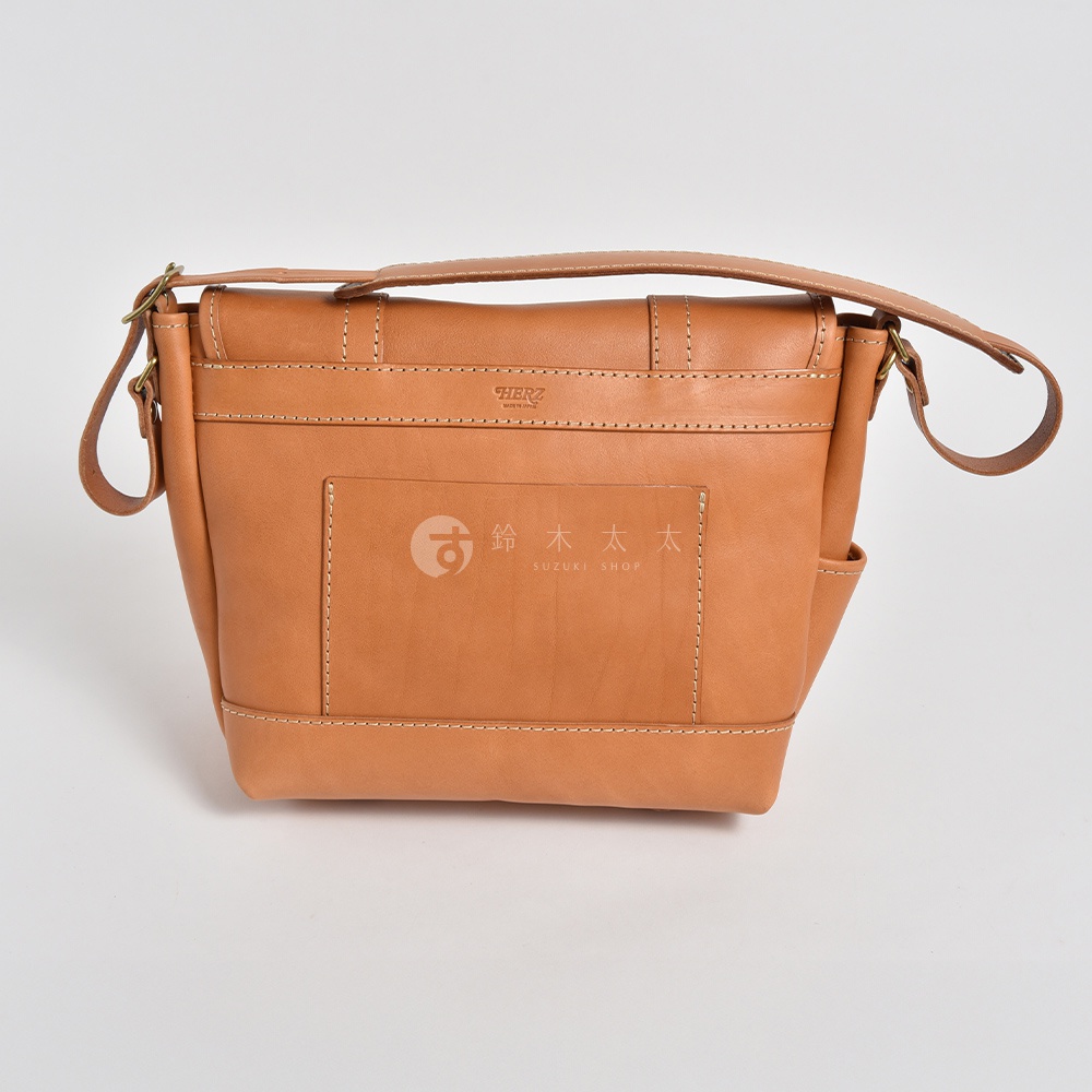 職人手作皮革復古郵差包，淺棕色，以全皮革製成，帶有明顯的縫線細節。包包前方有一個方形的皮革貼袋，上面印有品牌標誌及日文文字。包包附有可調節的肩帶，採用金屬扣環。