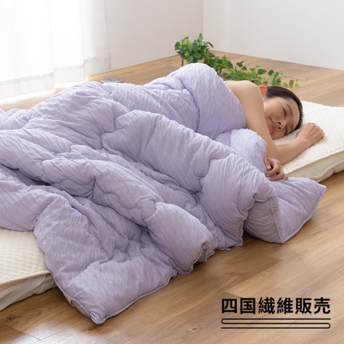 淺紫色舒芙蕾透氣四季被，中棉款，觸感蓬鬆柔軟，表面有細緻的紋理，覆蓋在一位女性身上，她正安詳地睡在床墊上，背景為木地板與植物。