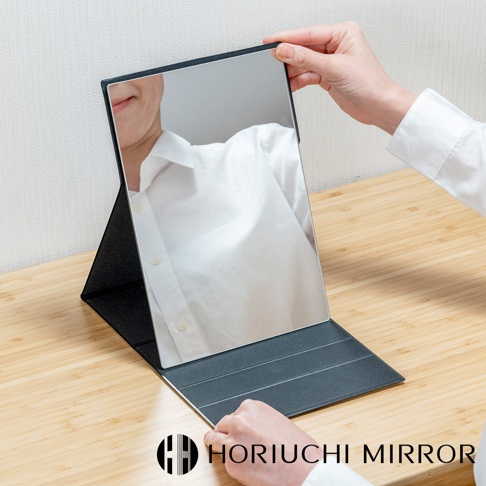 深灰色折疊式桌上化妝鏡，鏡面光滑，可調整角度，鏡框黑色，支架為深灰色，放在木紋桌面上。