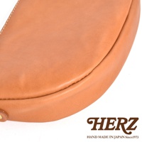 日本 HERZ 品牌皮革半圓馬鞍包特寫，淺棕色皮革質感，細膩縫線，底部有品牌 LOGO。
