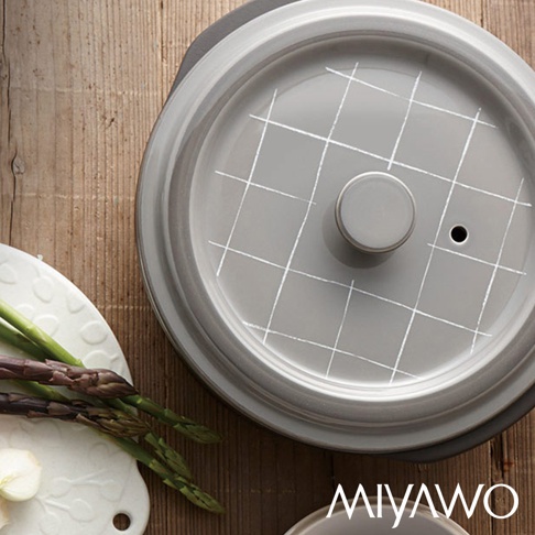 灰色的陶土湯鍋，鍋蓋上有白色網格線條裝飾，中央有圓形鍋鈕，鍋蓋一側有透氣孔。湯鍋置於木質桌面，旁邊有白色餐盤，盤上放著蘆筍和蒜頭。