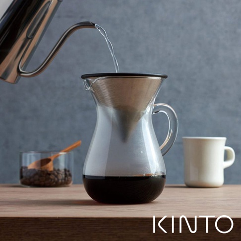 玻璃手沖咖啡壺組，包含一個透明玻璃咖啡壺，帶有銀色金屬濾網，上方有黑色邊緣。旁邊有一個白色馬克杯和一個裝有咖啡豆的玻璃杯，杯內有一支木勺。