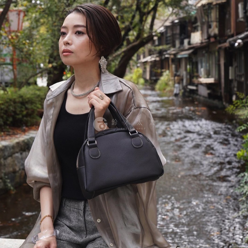 深灰色荔枝紋皮革製成的半月形迷你手提包，配有雙提把及拉鍊開口，由一位女性持握，她身穿一件淺灰色半透明外套和黑色上衣，背景為一條小溪與日式建築。