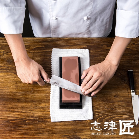 一名廚師正用磨刀石打磨一把刀，桌上還有一把未使用的刀，以及「志津匠 SHIZU TAKUMI」的標誌。