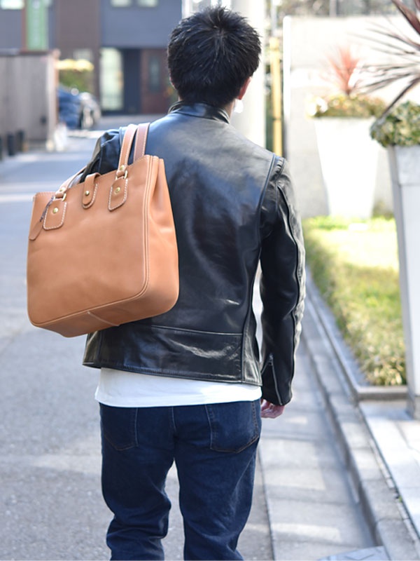一名男子身穿黑色皮衣，肩背棕色皮革托特包，包包上有原子釦設計。