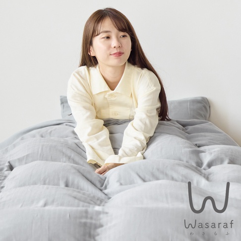 一位女性坐在淡黃色上衣，被灰色雙人床包及被套包圍，背景為白色牆壁，右下角有品牌 Logo。