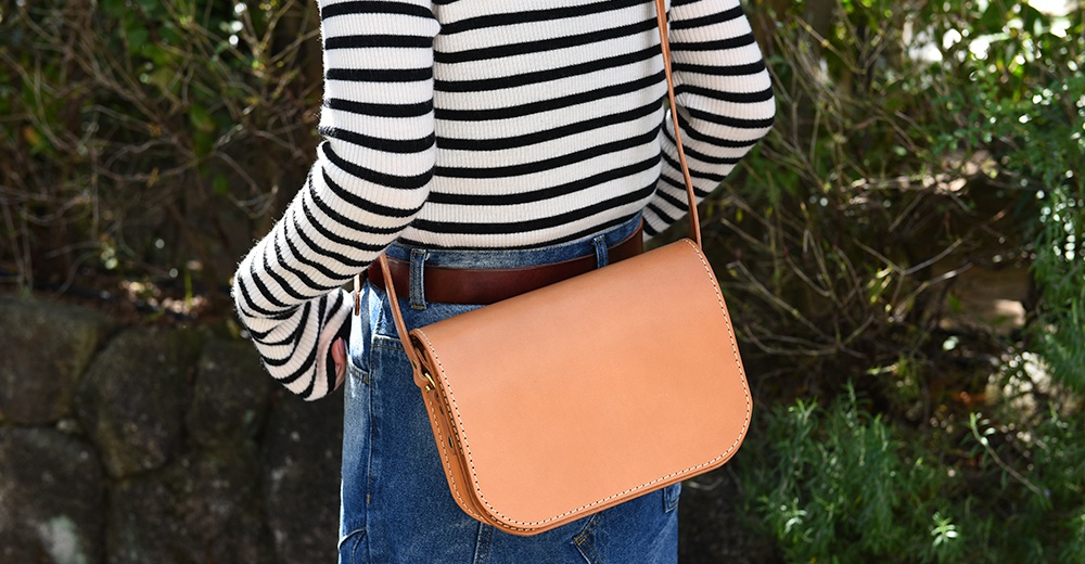淺棕色皮革翻蓋橫式單肩包，搭配黑色條紋上衣與牛仔褲，包身線條簡潔，縫線清晰可見。