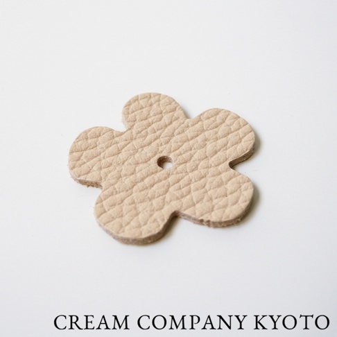 淺米色皮革製成的五瓣花形小掛飾，帶有壓紋紋理和中心孔洞。