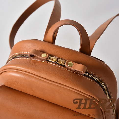 職人手作皮革立體口袋迷你後背包，採用棕色皮革，配有黃銅色拉鍊和鈕扣，以及清晰的白色縫線。