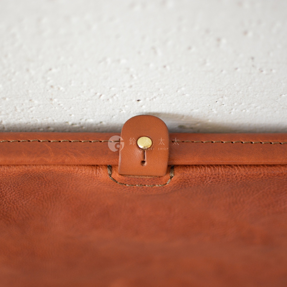 棕色皮革手拿包的局部特寫，展示了皮革的紋理、縫線細節和帶有黃銅按鈕的皮革搭扣。