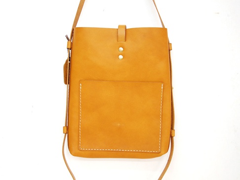 職人手作皮革散步直式單肩包，天然皮革材質，呈現溫潤的焦糖棕色。包身線條簡潔，正面設有一個方形貼袋，袋口以白色縫線勾勒，質感樸實。包蓋處有兩顆金屬卯釘，並以皮革搭扣固定。細長的皮革背帶垂掛，整體風格自然休閒。