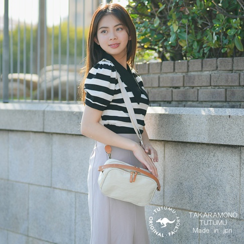 一位年輕女子拿著一個米白色、帶有棕色皮革飾邊的斜背包，她身穿黑白條紋上衣，站在戶外建築物旁。